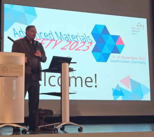 A person is standing on a stage behind a lectern that holds a laptop. In the background a presentation is visible on the screen with a conference logo for the conference "Advanced Materials Safety 2023" and "Welcome!"